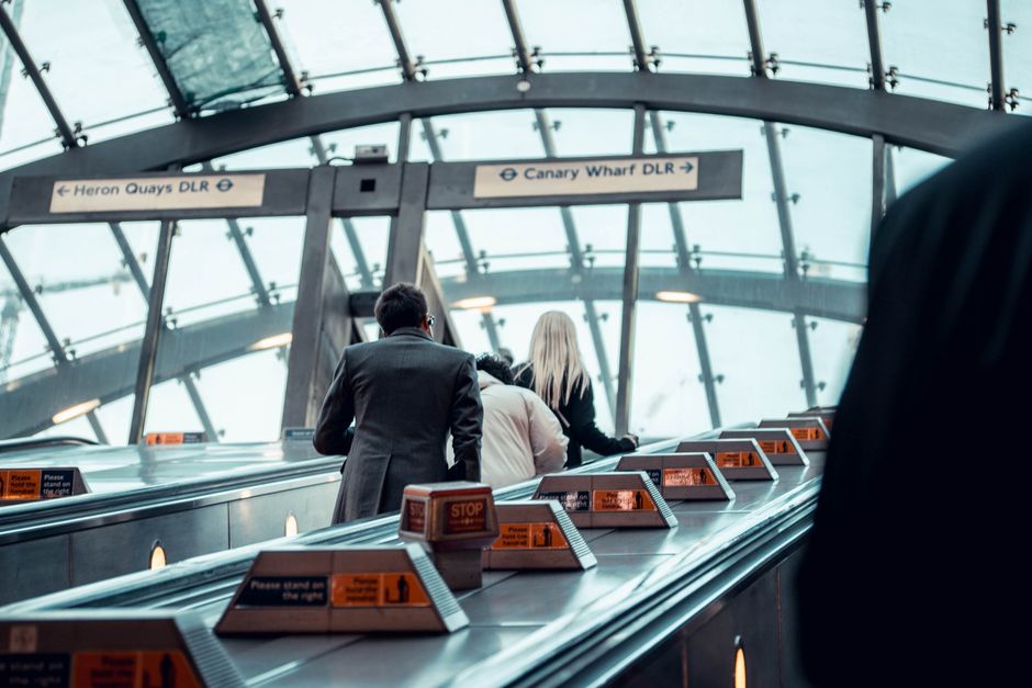 Inside Canary Wharf station, a high glass roof with a curved structure allows natural light to illuminate the area. In the foreground, a row of metal rubbish disposal units with orange warning and instruction labels is aligned along a counter or conveyor belt, likely for waste collection or sorting. Behind the units, three individuals are visible: two men and one woman, all dressed in business attire, positioned in the middle of the frame, appearing to be engaged in a task or conversation. The background features directional signs overhead, with one pointing to Heron Quays DLR and the other to Canary Wharf DLR, indicating the station's transit options. The environment is clean and modern, emphasizing efficient public space management. The presence of the rubbish bins suggests the station's facilities for maintaining cleanliness, possibly related to alternative waste handling or on-site collection efforts, supporting the context of private rubbish clearance and waste disposal services as part of the station’s maintenance routine.