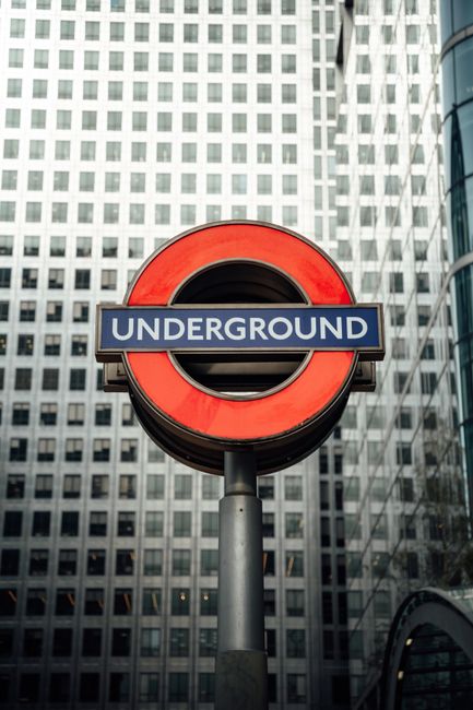 A prominent round London Underground sign featuring a red circle with a blue bar across the middle displaying the word 'UNDERGROUND' in white capital letters. The sign is mounted on a tall, cylindrical grey pole situated outdoors against a backdrop of modern high-rise office buildings with a grid-like pattern of numerous small, square windows. The buildings are made of light-colored materials and reflect a contemporary architectural style. The scene is illuminated by natural daylight, casting soft shadows and highlighting the vibrant colours of the underground sign. This urban environment, characterized by the densely packed structures and the iconic transit symbol, subtly relates to the topic of urban waste management and the importance of effective rubbish removal services in busy city areas, such as Canary Wharf. While only the sign is clearly visible, its placement suggests proximity to transportation hubs and shared city infrastructure, which can impact waste collection and disposal routines managed by companies like [COMPANY_NAME].