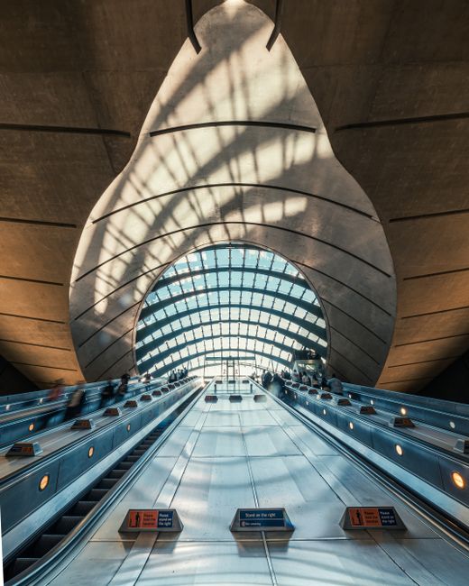 Inside Canary Wharf station, a high glass roof with a curved structure allows natural light to illuminate the area. In the foreground, a row of metal rubbish disposal units with orange warning and instruction labels is aligned along a counter or conveyor belt, likely for waste collection or sorting. Behind the units, three individuals are visible: two men and one woman, all dressed in business attire, positioned in the middle of the frame, appearing to be engaged in a task or conversation. The background features directional signs overhead, with one pointing to Heron Quays DLR and the other to Canary Wharf DLR, indicating the station's transit options. The environment is clean and modern, emphasizing efficient public space management. The presence of the rubbish bins suggests the station's facilities for maintaining cleanliness, possibly related to alternative waste handling or on-site collection efforts, supporting the context of private rubbish clearance and waste disposal services as part of the station’s maintenance routine.