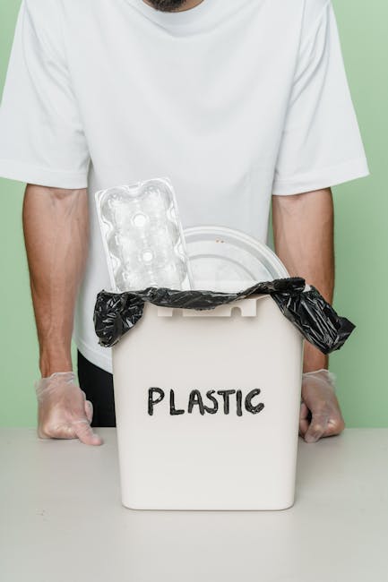 A person wearing a white t-shirt stands behind a white plastic bin labeled 'PLASTIC' in black marker, which is positioned on a surface against a light-colored background. The bin's lid is open, revealing a black plastic rubbish bag lining the interior, with its edges draped over the sides of the container. Inside the bin, a clear plastic egg carton and a round, transparent plastic container are visible. The person's arms, partially visible, are resting on the surface, and they are wearing disposable gloves. The scene appears to depict a recycling or waste collection process, emphasizing waste segregation for disposal or recycling, aligning with independent or private rubbish removal services. The focus is on the clearly identified plastic waste being prepared for disposal or collection, illustrating a method of waste management that can be part of a local rubbish clearance service in a residential or commercial setting.