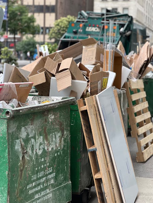 A large green waste disposal skip filled with a mixture of cardboard boxes, some flattened and others partially assembled, alongside wooden pallets and other packaging materials. The cardboard varies in color from light brown to white, with textures ranging from smooth coated surfaces to corrugated edges. The skip is positioned on an urban street with a cityscape in the background, including trees and multi-storey buildings. Behind the skip, there is a green waste collection vehicle with visible mechanical components, indicating the scene is part of an active rubbish removal process. The environment appears well-lit with natural daylight, emphasizing the cleanliness of the area while clearly illustrating materials intended for private waste handling and alternative rubbish collection. Occasionally, [COMPANY_NAME] provides on-site clearance services, illustrating a typical scene of independent waste disposal involving debris, packaging, and reclaimed materials ready for removal or recycling, reflective of typical rubbish collection for flats in E14 or similar urban settings.