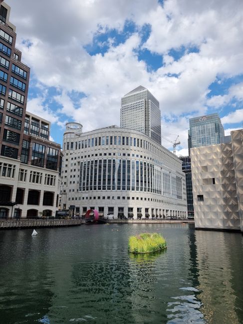 The image depicts a cityscape view of Canary Wharf with a focus on a modern, white, multi-story office building situated along a waterway. The building features a distinctive curved facade with horizontal bands of windows and a textured surface, giving it a sleek, contemporary appearance. To the right, partially visible, is a structure with a geometric, diamond-patterned design in a metallic finish, adding architectural variety to the scene. Behind the main building, two larger office towers are visible, one with a glass facade reflecting the sky and nearby structures, and the other with a more solid, grid-patterned exterior. The water in the foreground appears calm, with minimal ripples, and contains a small, irregularly shaped patch of green foliage floating near the centre. The sky above is partly cloudy with patches of blue, casting natural light that highlights the buildings' surfaces. The entire scene has a professional, urban atmosphere consistent with a central business district, where such structures support various commercial activities, including independent office rubbish removal that organisations like [COMPANY_NAME] might facilitate as part of their waste management solutions.