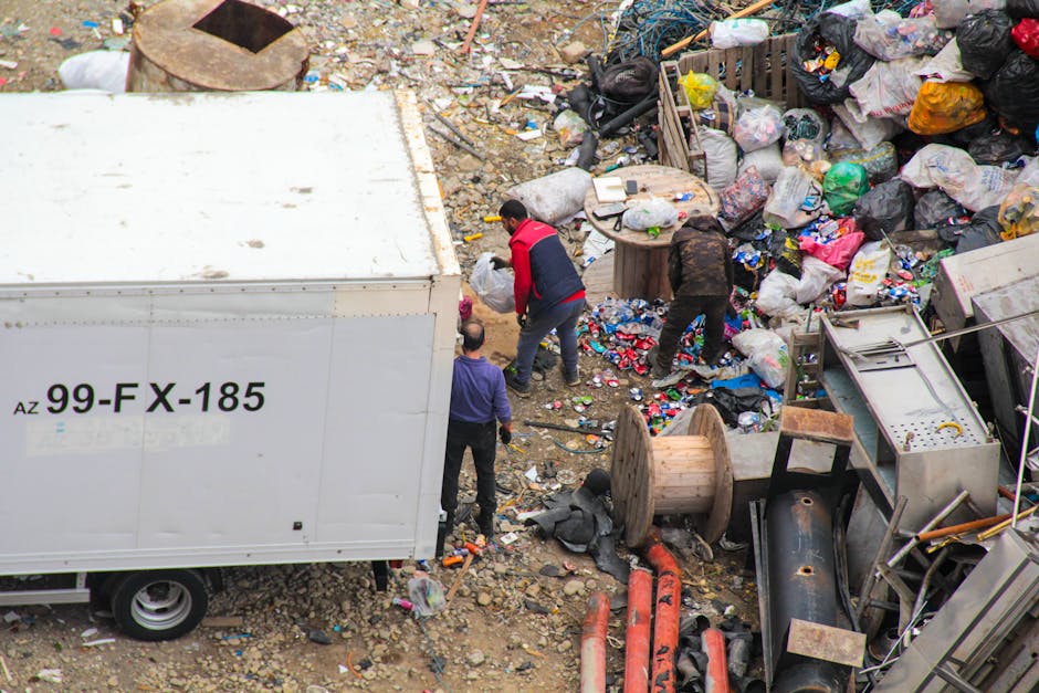 An outdoor scene showing a large amount of mixed household waste and debris scattered across a gravel surface, with several individuals actively sorting and removing rubbish around a white enclosed trailer marked with the code 'AZ 99-FX-185'. The waste includes numerous black, white, and multicoloured plastic bags filled with discarded items, along with loose litter such as paper, packaging, and small debris. The background features a cluttered area with wooden spools, pipes, and various discarded appliances or machinery parts, partially obscured by the waste. The lighting is natural, suggesting daytime, and the environment appears to be an open space used for waste collection or clearance activities, aligning with private rubbish removal services like those offered by Rubbish Clearance Canary Wharf. The scene emphasizes hands-on waste management and ongoing rubbish clearance efforts in a non-residential setting.
