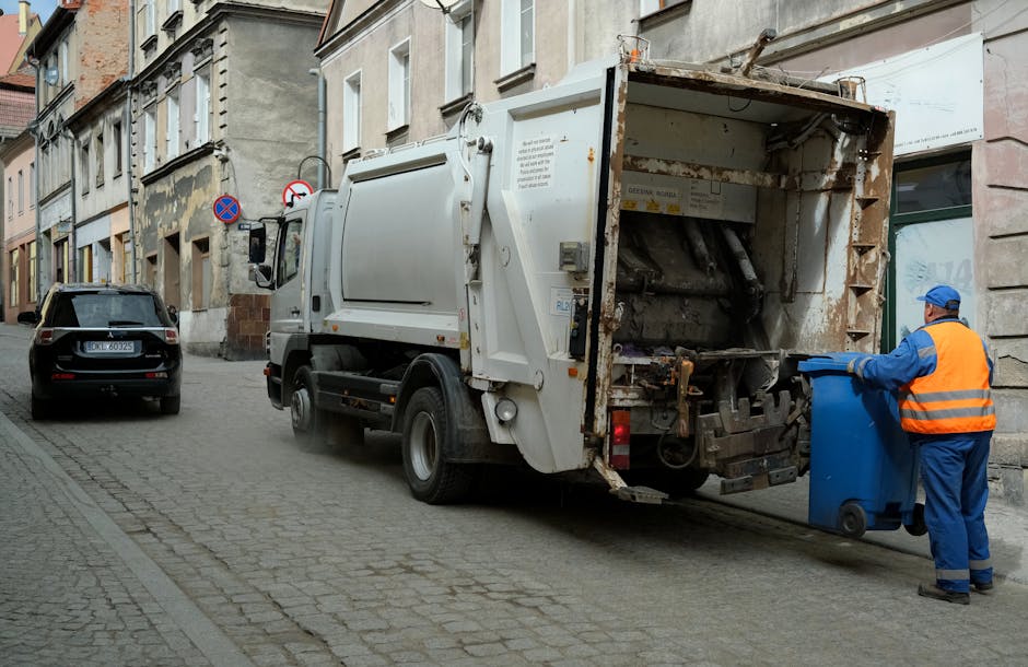 A black vehicle parked on a narrow cobblestone street adjacent to old, multi-story buildings with weathered facades. The street scene includes a large white rubbish collection truck with rust and dirt visible on its rear compartment, positioned close to the sidewalk. A worker dressed in a blue uniform and orange high-visibility vest is standing beside the truck, emptying a blue wheeled bin into the rear opening of the vehicle. The worker wears a blue cap and gloves, and is focused on the task. The background shows additional parked cars and signage indicating no parking zones, with natural daylight illuminating the scene. The environment suggests typical urban waste collection in a historic area, reflecting private or independent rubbish removal efforts aligned with local waste management practices.