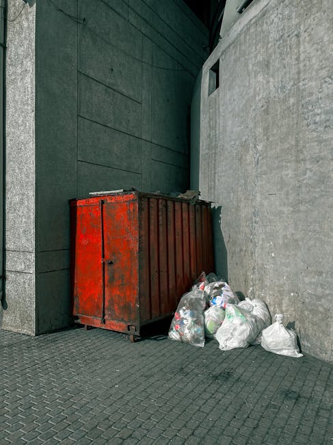 An outdoor scene showing a large amount of mixed household waste and debris scattered across a gravel surface, with several individuals actively sorting and removing rubbish around a white enclosed trailer marked with the code 'AZ 99-FX-185'. The waste includes numerous black, white, and multicoloured plastic bags filled with discarded items, along with loose litter such as paper, packaging, and small debris. The background features a cluttered area with wooden spools, pipes, and various discarded appliances or machinery parts, partially obscured by the waste. The lighting is natural, suggesting daytime, and the environment appears to be an open space used for waste collection or clearance activities, aligning with private rubbish removal services like those offered by Rubbish Clearance Canary Wharf. The scene emphasizes hands-on waste management and ongoing rubbish clearance efforts in a non-residential setting.